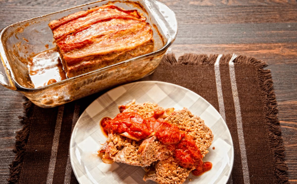 bacon and marinara meatloaf slice on white and brown plate in front of glass loaf pan sitting on brown placemat
