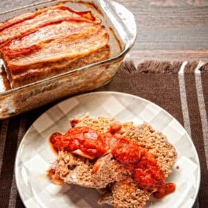 two slices of meatloaf on white checked plate with sauce on top next to glass loaf pan of meatloaf