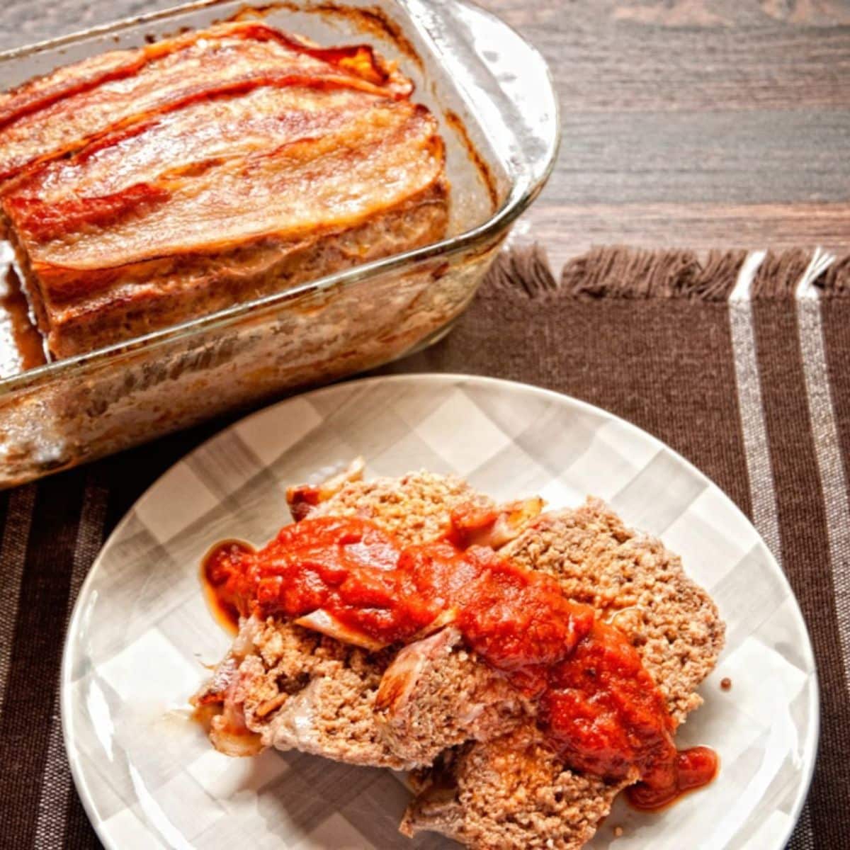 two slices of meatloaf on white checked plate with sauce on top next to glass loaf pan of meatloaf