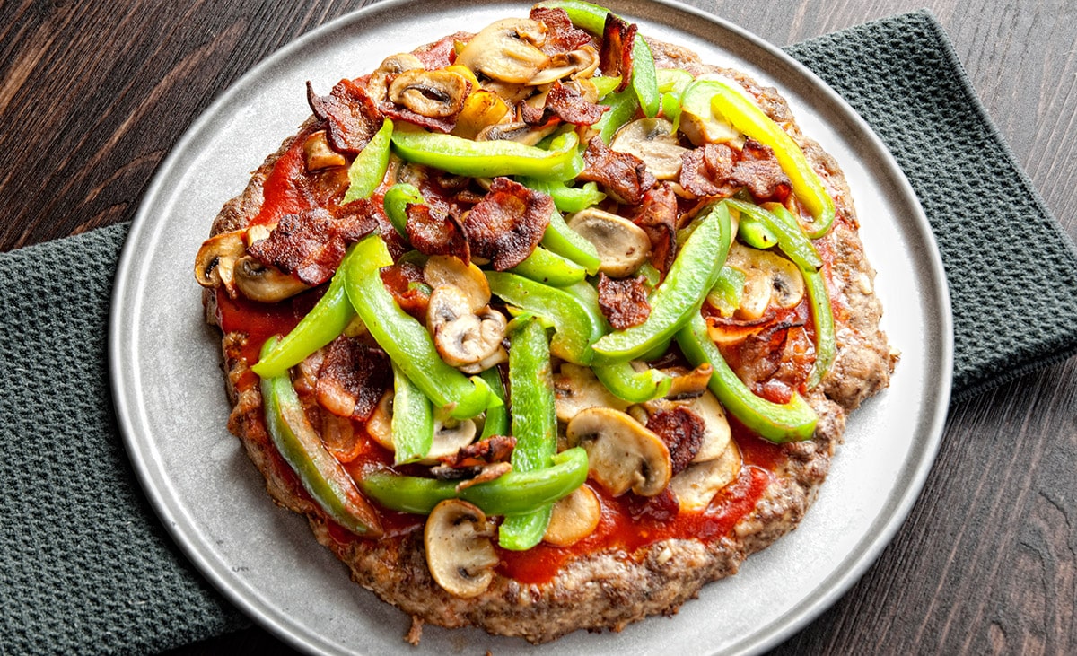 white plate with small meat pizza topped by mushrooms bell peppers and bacon