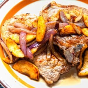 white plate with orange rim holding pork chops topped with apples and onions that are caramelized