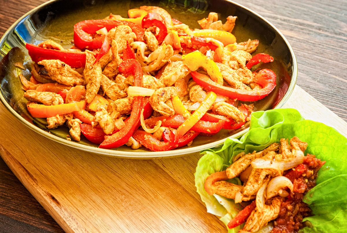 skillet of chicken fajitas with red bell peppers on wood board next to leaf lettuce filled with chicken