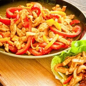 skillet of chicken fajitas with red bell peppers on wood board next to leaf lettuce filled with chicken