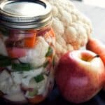 Jar of Pickled Vegetables with a head of raw cauliflower and an apple on a table