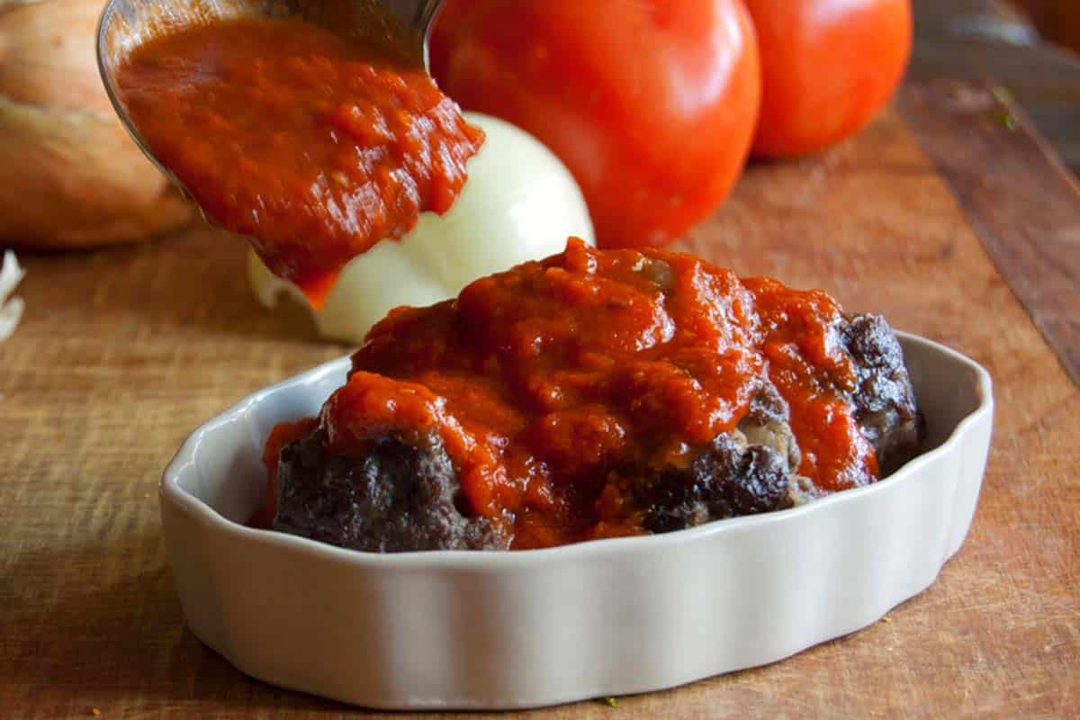 homemade Marinara sauce being spooned onto meatballs in a white ramekin dish