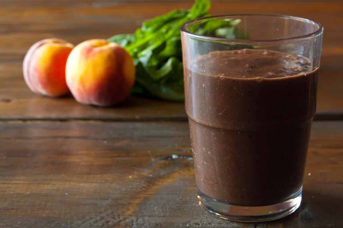 glass of Peach and chocolate green smoothie on a wood table with peaches in the background