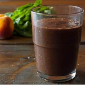 glass of Peach and chocolate green smoothie on a wood table with peaches in the background