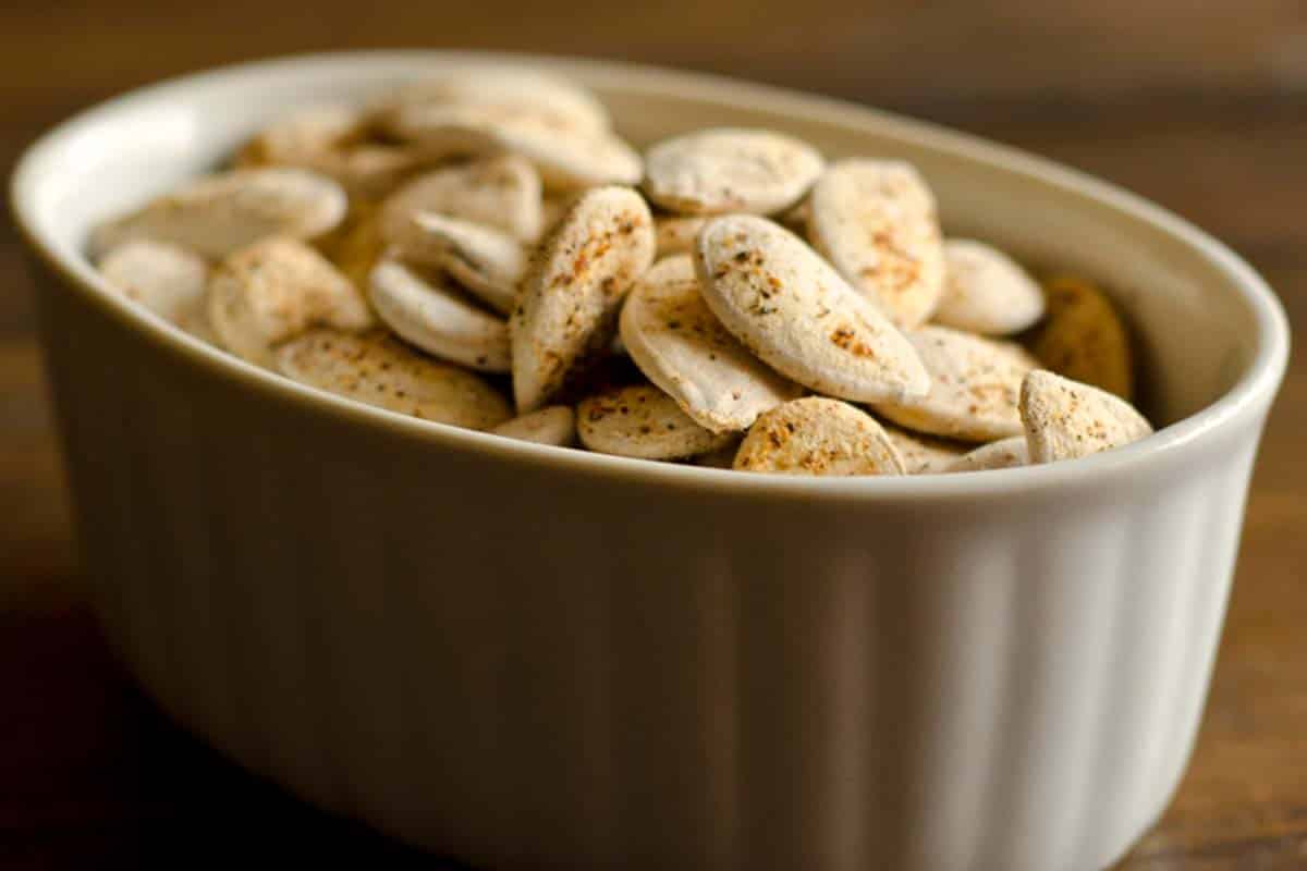 closeup of a white bowl filled with Spicy Pumpkin Seeds on a wood table