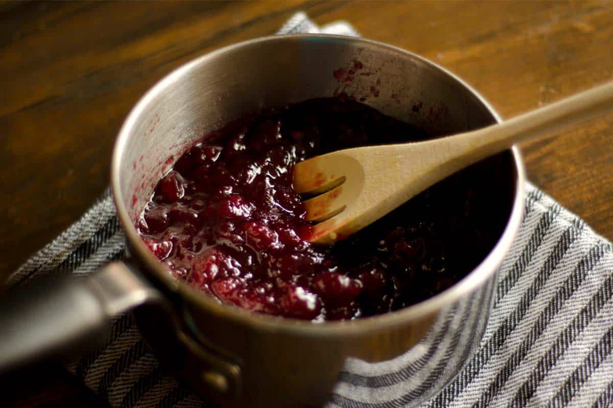 Tart cranberry sauce being stirred by a wood spoon in a saucepan on a table 